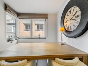 A dining area with a wooden table, two windows with curtains, a wall clock, and a lamp. - Luxury 4-bedroom apartment with large terrace (Blankenberge)