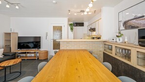 A modern living room with a wooden dining table, grey chairs, a flat-screen TV, a bookshelf, and a kitchen area with a brick backsplash. - Spacious Duplex Apartment in Blankenberge (Blankenberge)