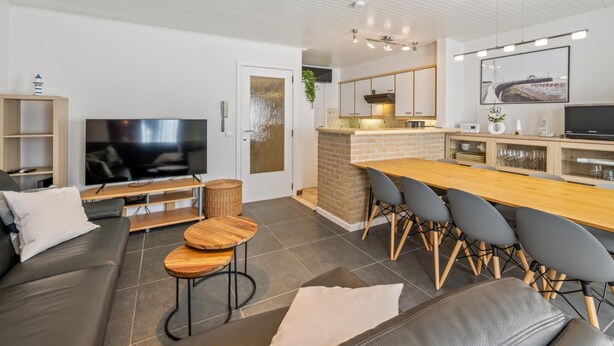 A modern living room with a flat-screen TV, a sofa, a dining table with chairs, and a kitchen area with a stove and cabinets. - Spacious Duplex Apartment in Blankenberge (Blankenberge)