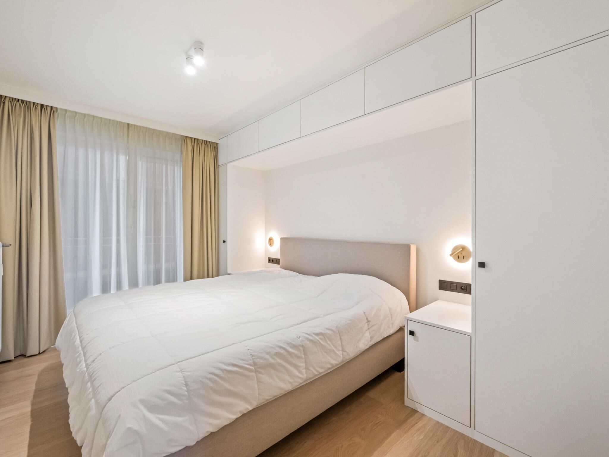 A modern hotel room with a large bed, built-in wardrobe, and a large window with curtains.