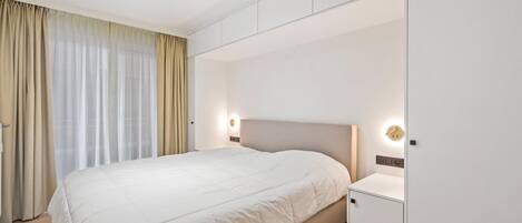 A modern hotel room with a large bed, built-in wardrobe, and a large window with curtains.