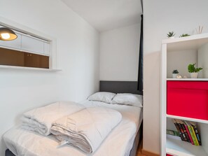 Ein ordentlich bezogenes Bett mit weißer Decke, ein Nachttischregal mit Büchern und eine kleine Pflanze auf dem Regal.