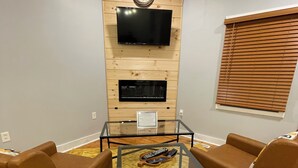 Un salon avec une table basse en verre, deux canapés en cuir marron, un téléviseur à écran plat fixé à un mur en bois, et une cheminée située sous le téléviseur.