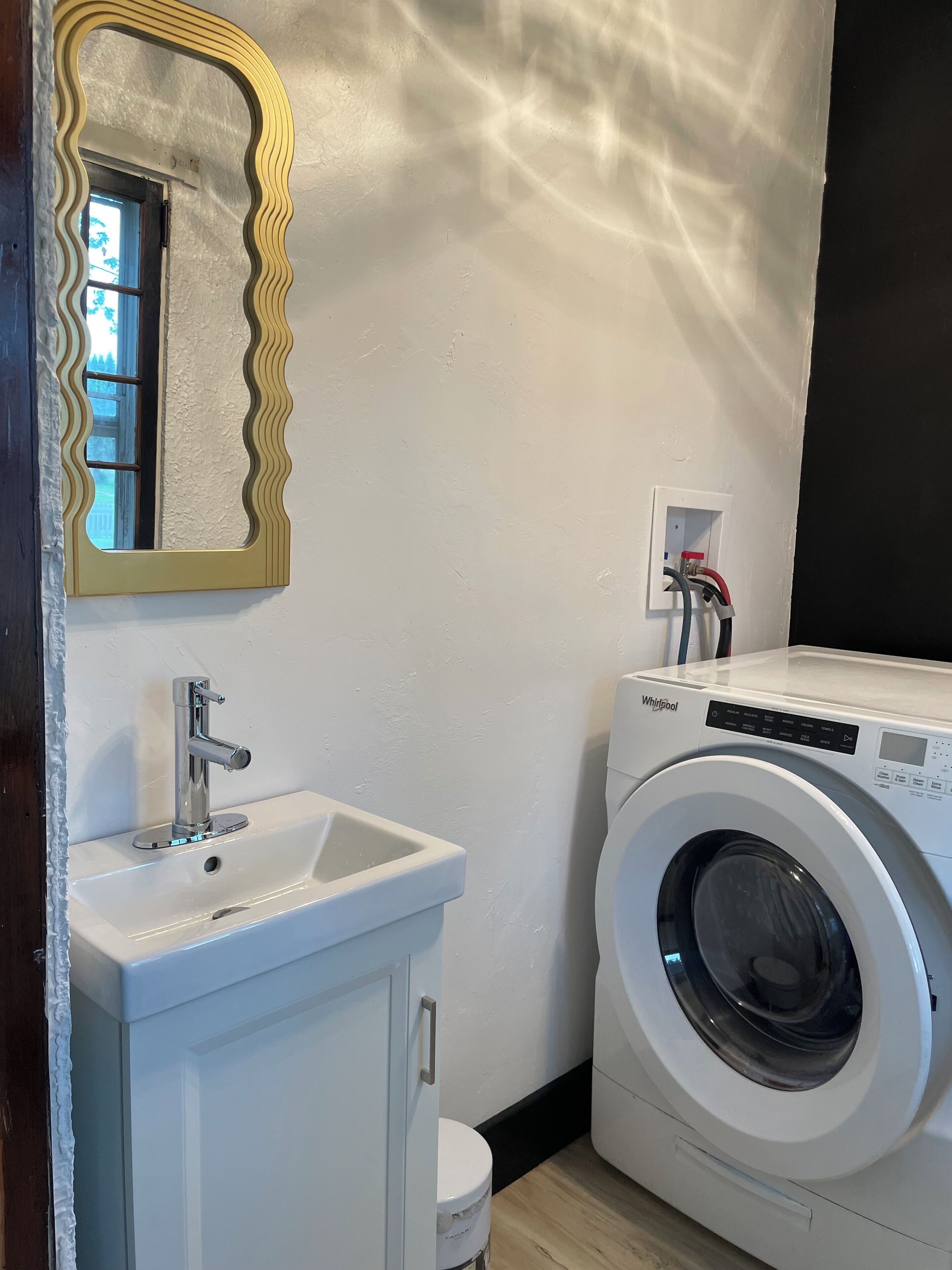 A compact bathroom with a white sink, a mirror with a wavy frame, a washing machine, and a power outlet.