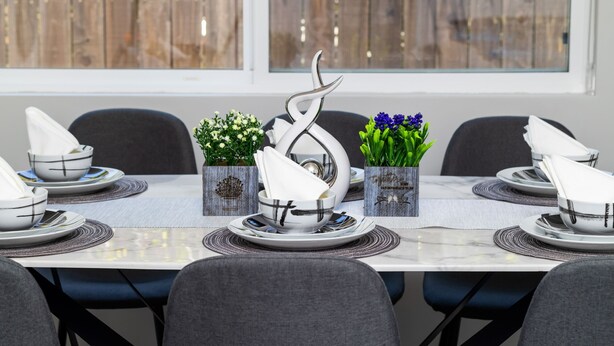 Una mesa de comedor dispuesta con platos, tazones y servilletas blancos, flanqueada por jarrones decorativos con flores.