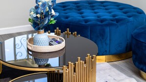 Una habitación moderna con una mesa redonda de vidrio, un otoman de terciopelo azul y un jarrón decorativo con flores.