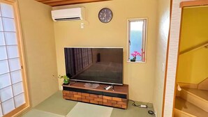 A room with tatami flooring, a television on a wooden stand, a clock on the wall, and a small window with a plant. - SUN WARM HOUSE (Osaka)