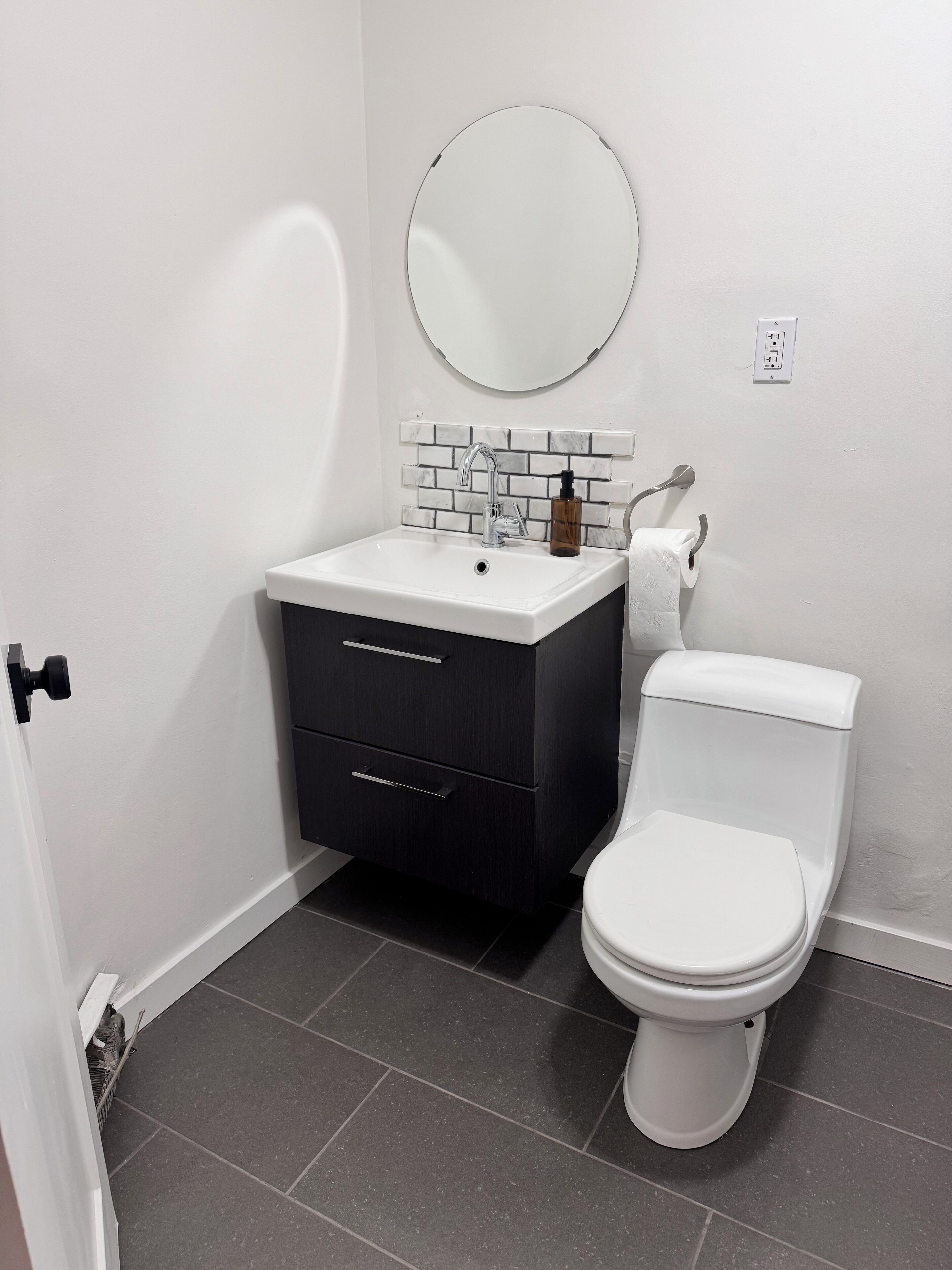 A bathroom with a white sink, dark cabinet, round mirror, and white toilet.