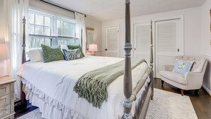A four-poster bed with a green throw blanket, white bedding, and multiple pillows. There is a chair with a pillow, a nightstand with a lamp, and a window with curtains. - Close to Connecticut College & Coast Guard Academy (Quaker Hill)