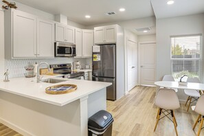 Uma cozinha moderna com armários brancos, eletrodomésticos de aço inoxidável, uma ilha central com banquetas e uma área de refeições com mesa e cadeiras brancas.