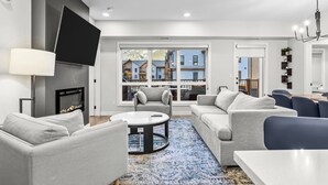 A modern living room with a fireplace, grey sofas, a coffee table, and a large window offering a view of neighboring buildings. - 3 bedroom + 2 bath -🏔️Rockies Refuge⛰️ heated outdoor pool and hot tub/full gym (Dead Man's Flats)