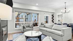 A modern living room with a fireplace, grey sofas, a round coffee table, and large windows offering a view of a blue house. - 3 bedroom + 2 bath -🏔️Rockies Refuge⛰️ heated outdoor pool and hot tub/full gym (Dead Man's Flats)