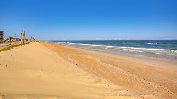 Ein Strand mit blauem Himmel, sandigem Ufer und Gebäuden im Hintergrund.