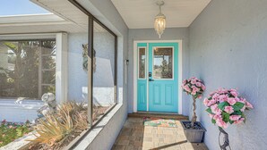 Une véranda couverte avec une porte bleue, des plantes en pot et un sol carrelé.