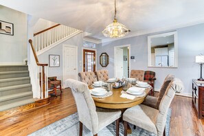 Un coin repas avec une table ronde dressée pour six personnes, entourée de chaises rembourrées. Un escalier avec des rampes en bois est visible sur la gauche.