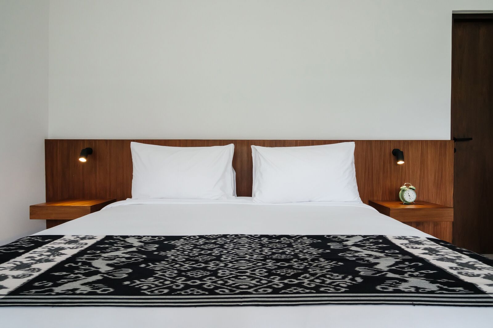 Un lit avec une couvre-lit à motifs, une tête de lit en bois, deux oreillers blancs, des tables de chevet avec des lampes et un petit réveil vert.