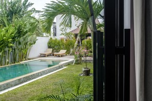 A swimming pool with lounge chairs, surrounded by lush greenery and a white building with a tiled roof. - Sunset Loft Seminyak, Bali (Seminyak)