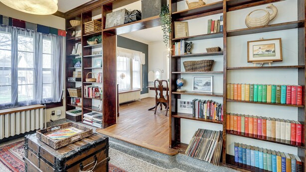 A room with wooden bookshelves filled with books and decorative items, a vintage trunk, and a globe on a table. - Historic Home Overlooking Central Riverside Park! (Wichita)