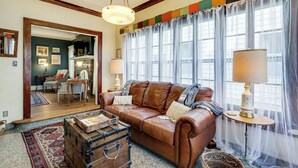 A living room with a brown leather sofa, a wooden coffee table, and a lamp. There is a large window with sheer curtains and a view of the outside. - Historic Home Overlooking Central Riverside Park! (Wichita)
