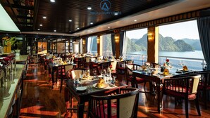 A restaurant with multiple dining tables set for guests, large windows offering a view of a body of water and hills, and a ceiling with a logo. - Aqua Luxury Cruise (Tuan Chau)