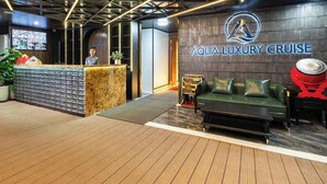A reception area with a wooden reception desk, a green sofa, and a wall with the logo 'Aqua-Luxury Cruise'. - Aqua Luxury Cruise (Tuan Chau)