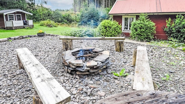 Un espace extérieur rustique aménagé autour d’un foyer, avec un banc en bois, le tout entouré d’un sol rocheux et avec un bâtiment rouge en arrière-plan.