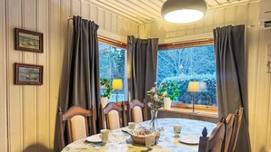 Un coin repas avec une table ronde dressée pour quatre personnes, entourée de chaises en bois. Deux tableaux encadrés ornent le mur, il y a une fenêtre avec des rideaux et une lampe sur l’appui de fenêtre.