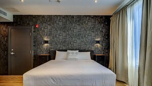 A modern bedroom with a large bed, a dark wooden door, a patterned wall, and a window with curtains. - Dream Suites - Deluxe King Suite (MISSISSAUGA)