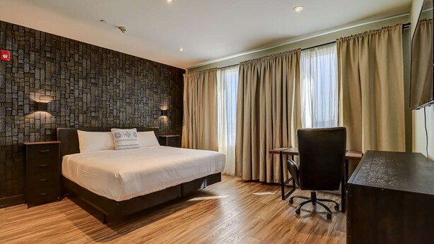 A hotel room with a large bed, a desk with a chair, a dark patterned wall, and a window with curtains. - Dream Suites - Deluxe King Suite (MISSISSAUGA)