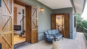 Uma área externa coberta com um sofá e uma pequena mesa de madeira, portas de madeira que dão para um espaço interior e um luminário na parede.