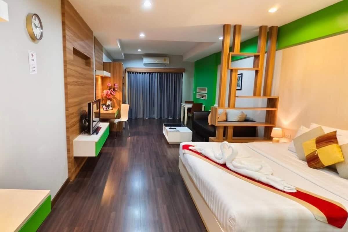 A hotel room with a bed, bedside table, and a desk with a chair. There is a window with curtains, a clock on the wall, and an air conditioning unit.