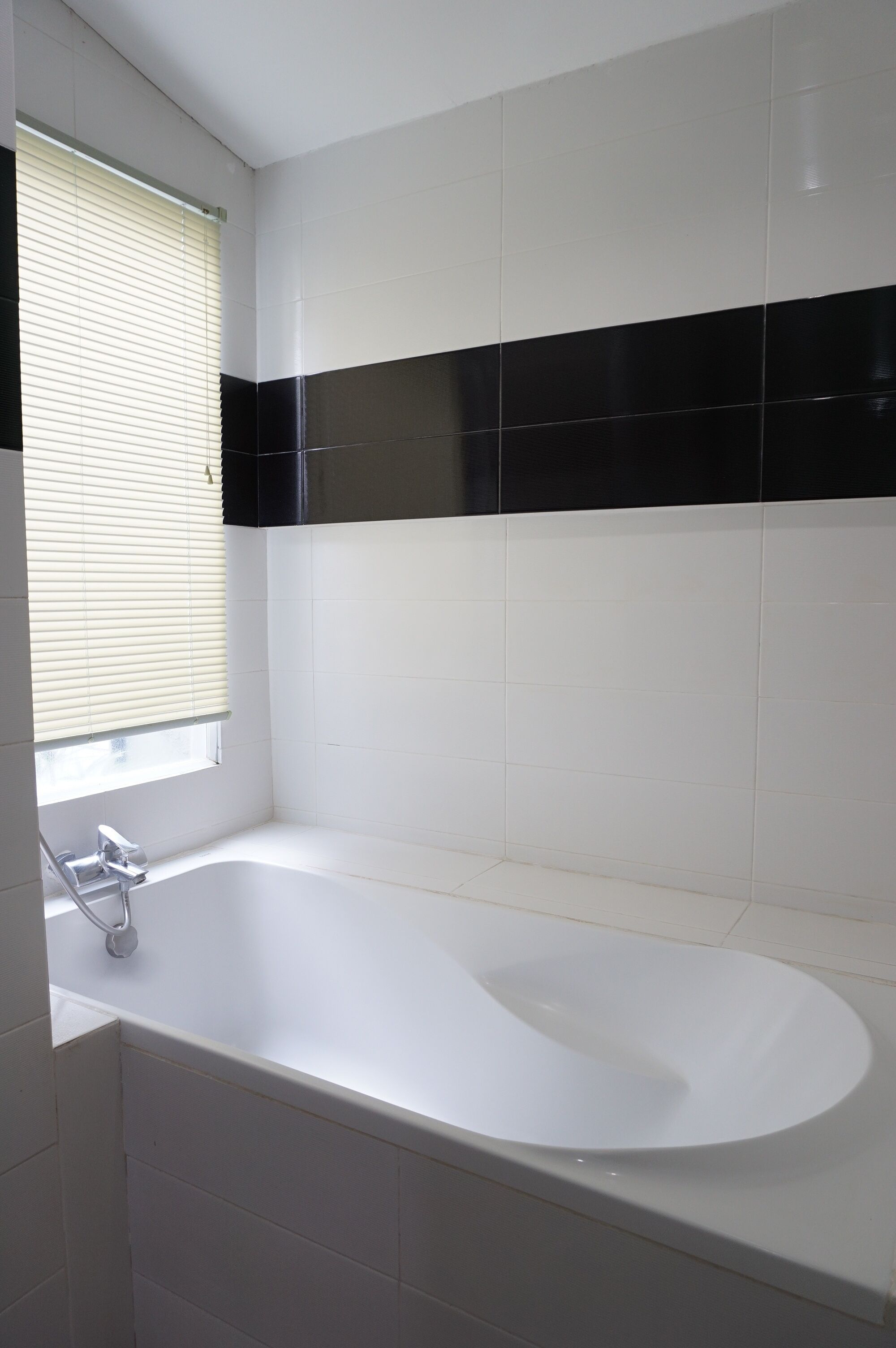 A bathroom with a bathtub, a window with blinds, and black and white tiles.