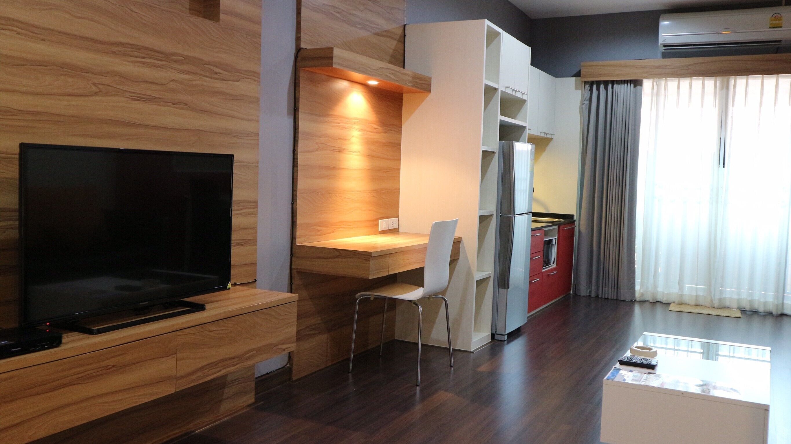 Una sala de estar moderna con un televisor de pantalla plana, muebles de madera, un banco empotrado y una zona de cocina con refrigerador y armarios.