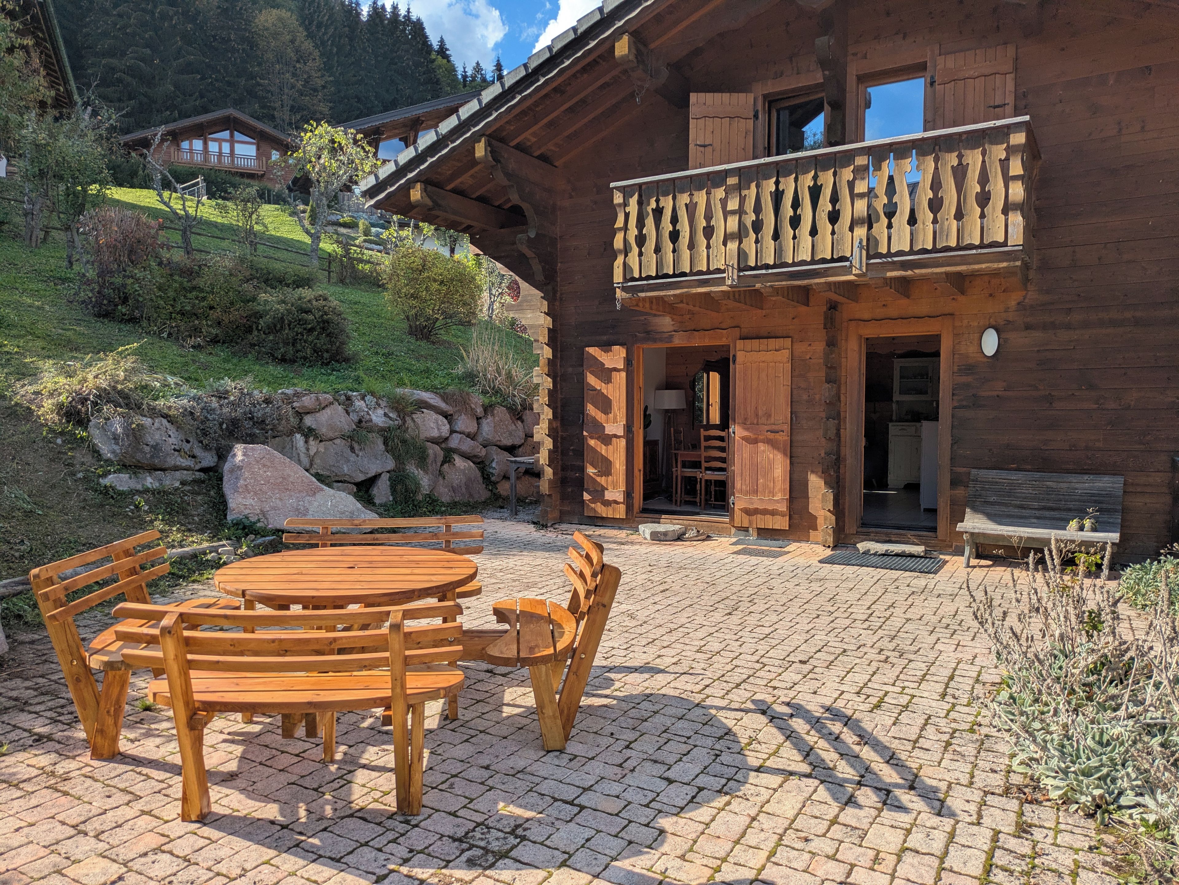 Un chalet en bois doté d’un balcon, d’un mur en pierre et d’une terrasse dallée avec des meubles en bois.