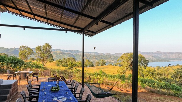 An outdoor dining area with a table set for four, surrounded by chairs, under a covered patio with a view of a lake and hills. - Cosmicstays Birdsong Lakeview Cottage  (Velhe)