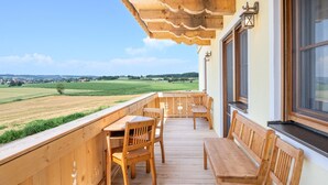 Een houten balkon met een tafel en stoelen, een bankje en uitzicht op een groen veld en gebouwen in de verte.