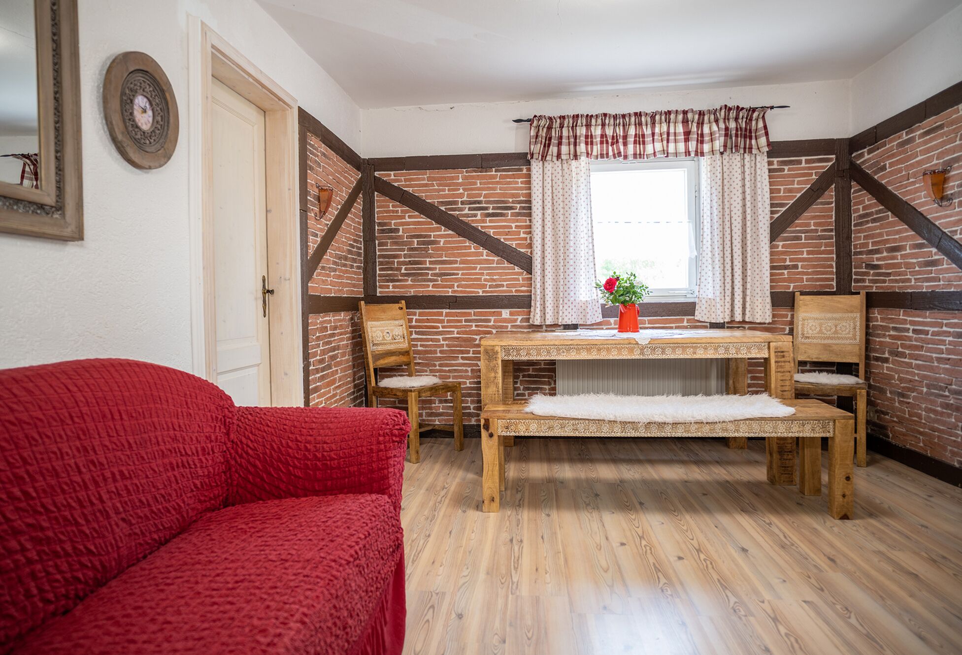 Ein gemütliches Zimmer mit Holztisch, zwei Stühlen, einer roten Couch und einem Fenster mit Vorhängen.
