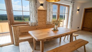 Una mesa de madera con bancos, un florero con plantas secas y vistas al campo a través de amplios ventanales.
