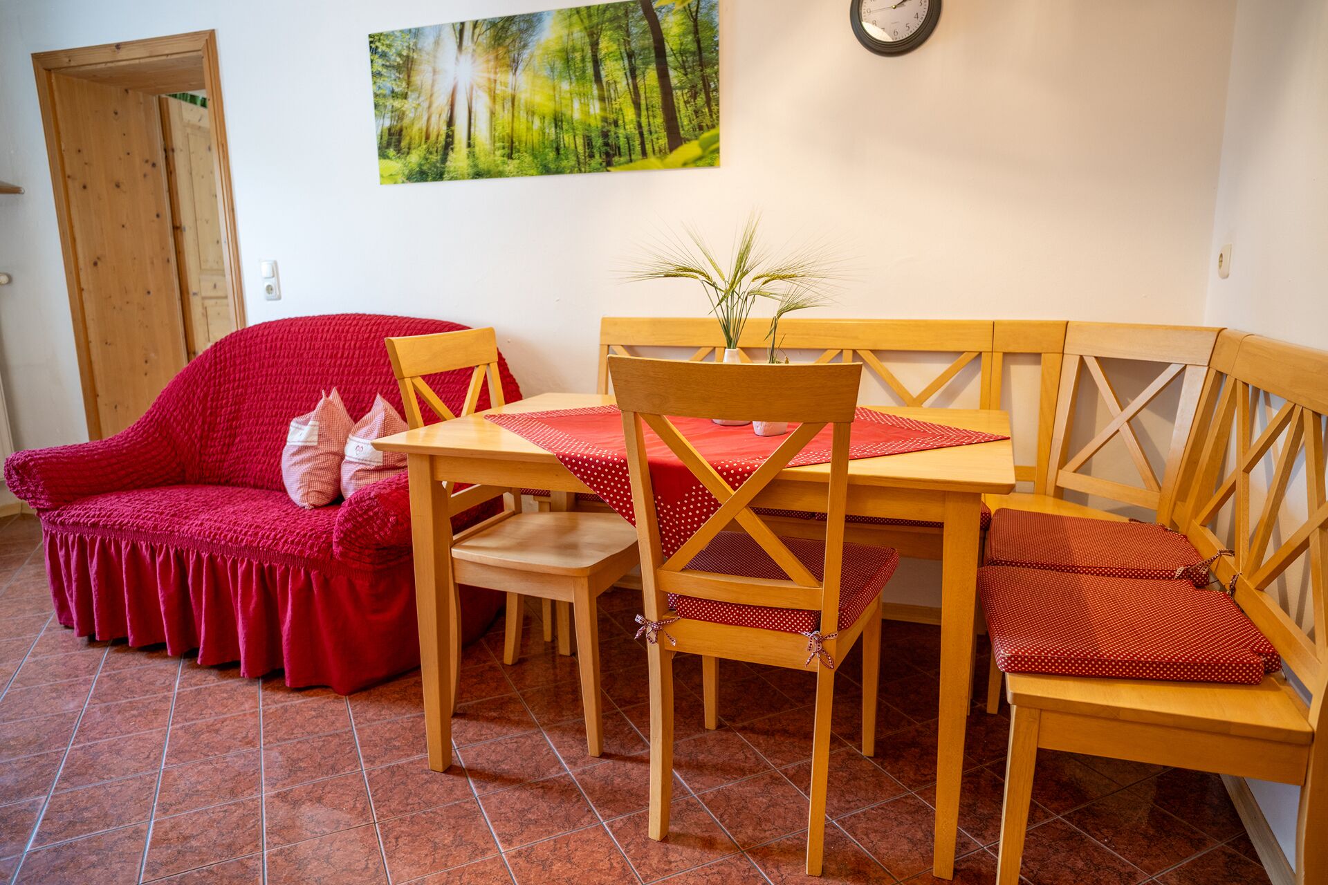 Une pièce chaleureuse avec un canapé rouge, une table à manger en bois et des chaises. Un tableau représentant une forêt est accroché au mur, et une horloge est visible.