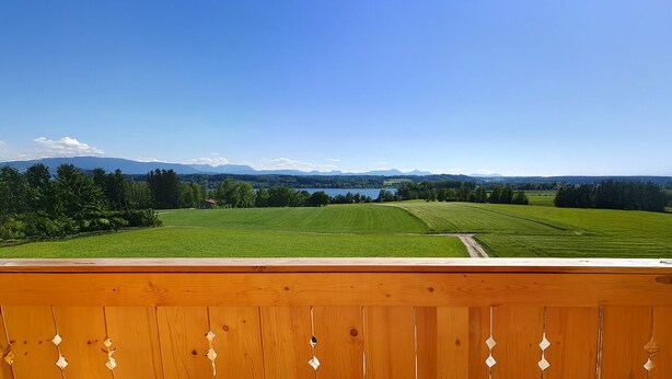 Udsigt fra en balkon over et grønt marker, en sø og fjerne bjerge.
