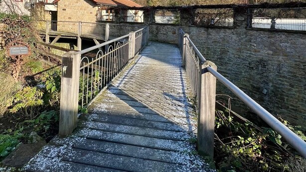 Un pont de pierre avec des garde-corps métalliques menant à un bâtiment en pierre surmonté d’un toit rouge.