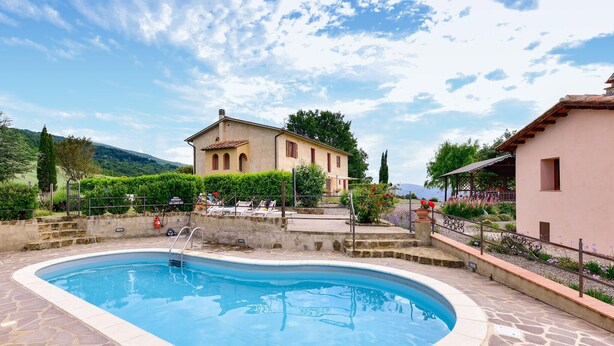 Une piscine aux bords recouverts de pierres, un bâtiment doté d’un toit en tuiles rouges et un jardin agrémenté de fleurs et d’arbustes.