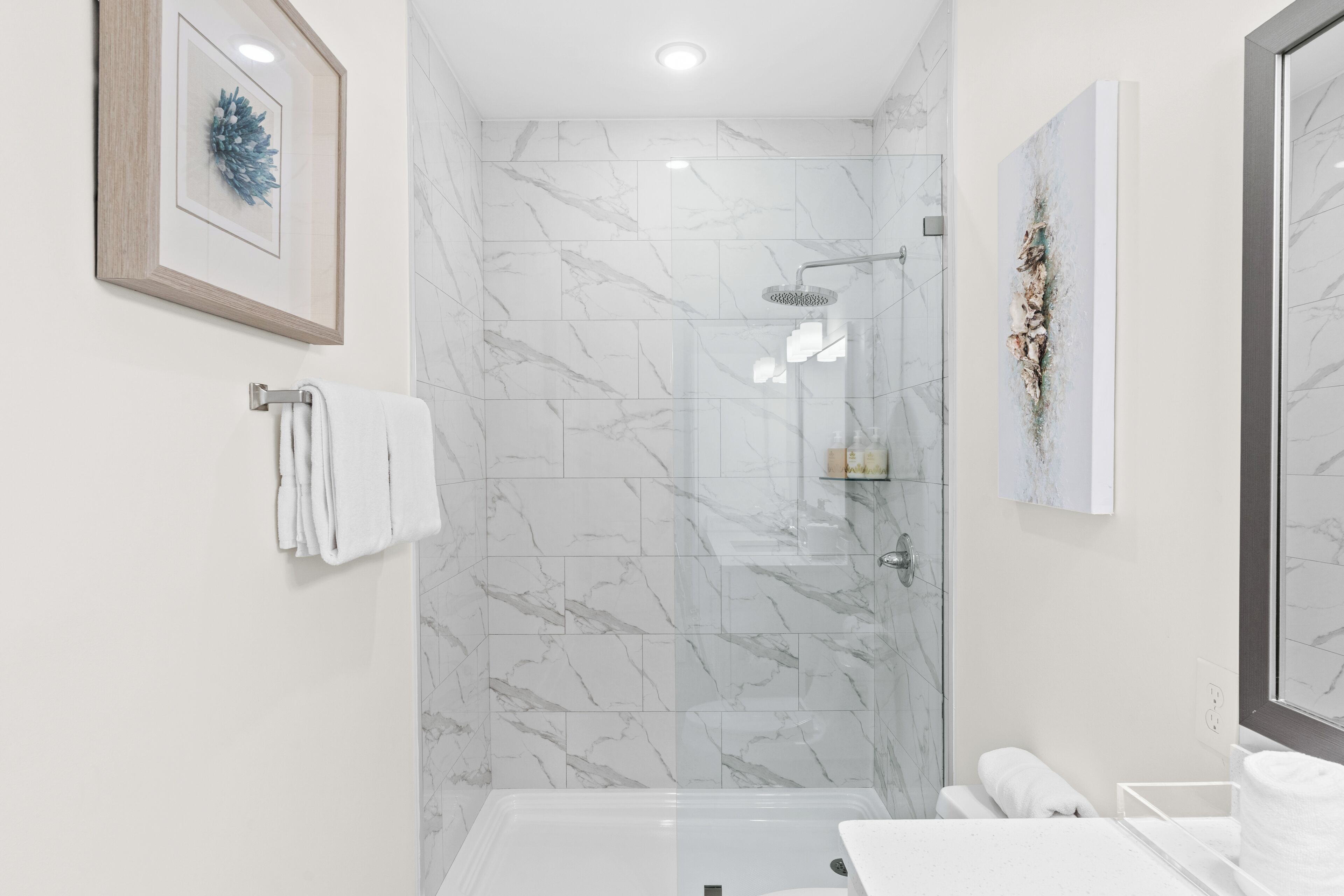 Un baño moderno con ducha envuelta en vidrio, azulejos de mármol y dos obras de arte enmarcadas en las paredes.