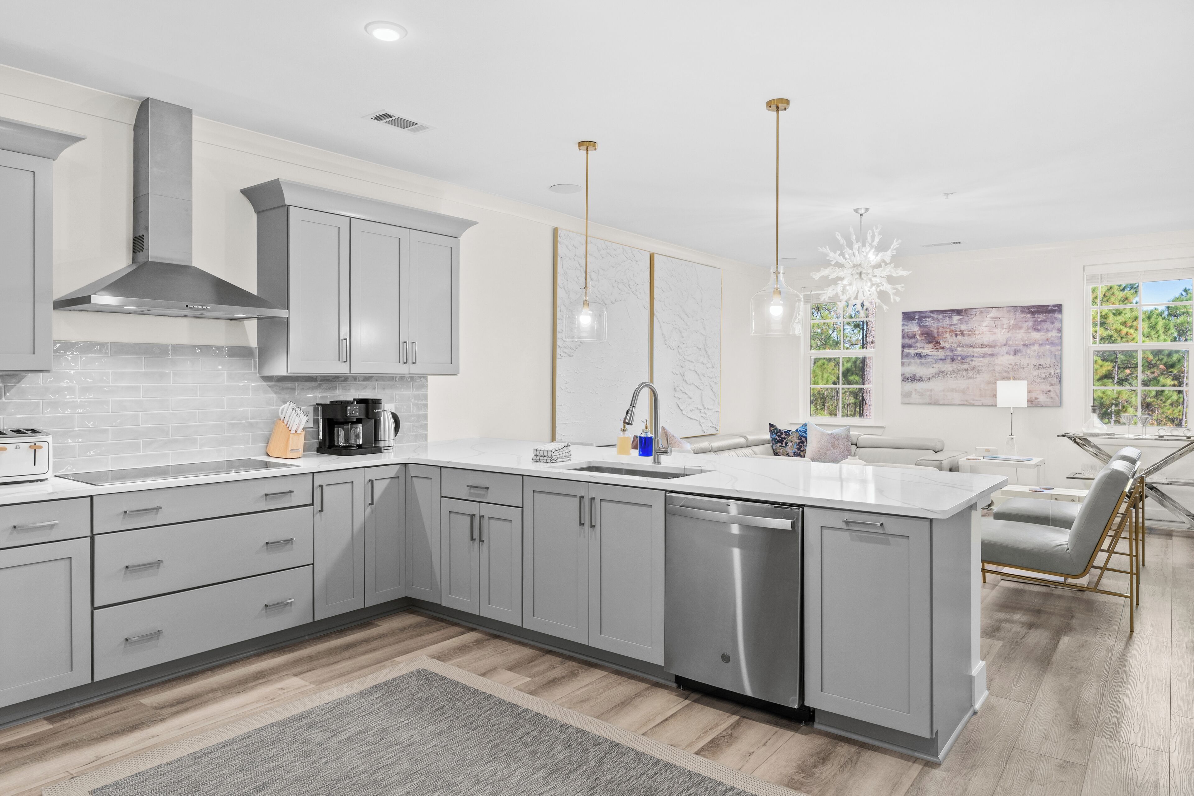A modern kitchen with grey cabinets, a dishwasher, and a large island. There are pendant lights and a view of the outdoors through large windows.