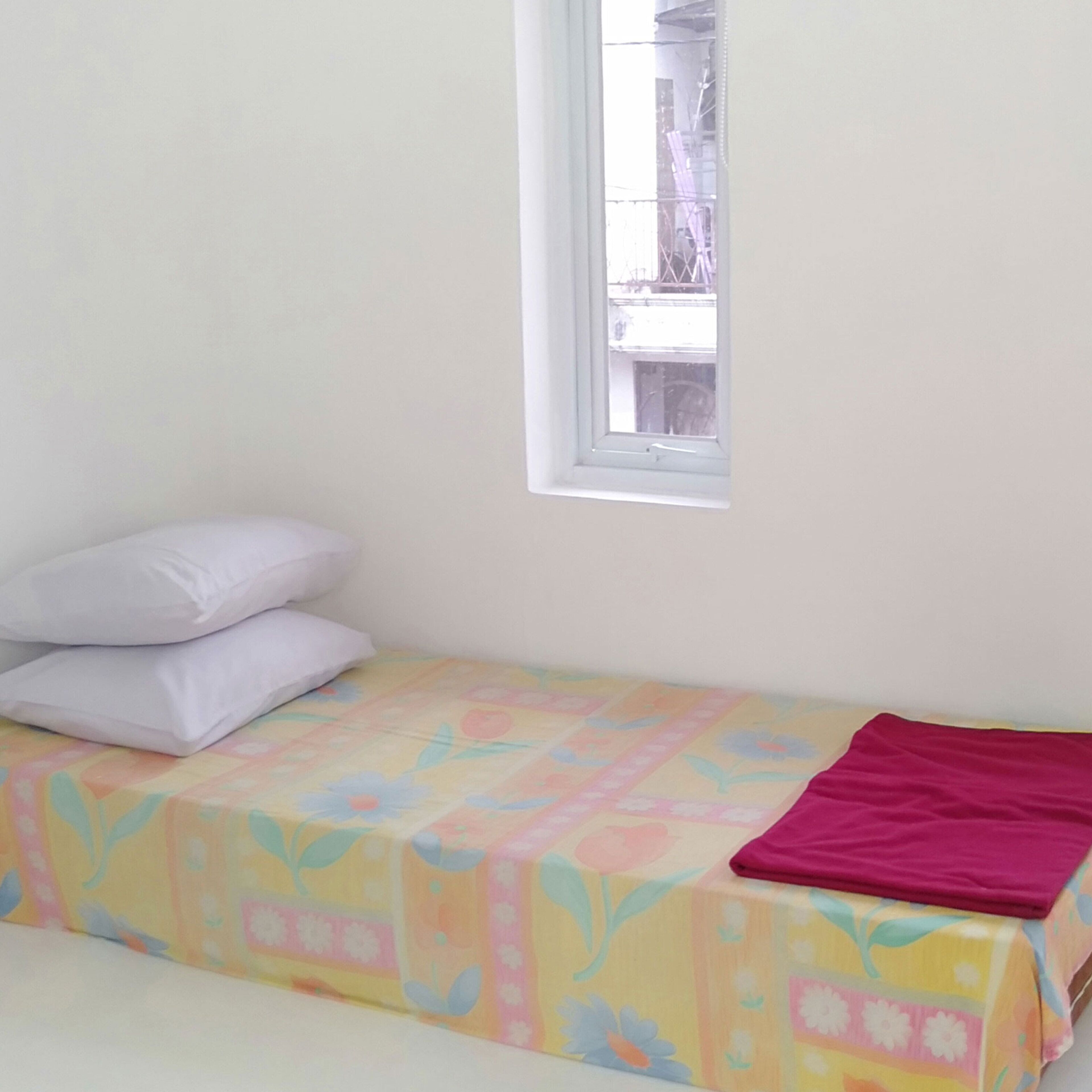 A small, single-bed room with a colorful bedspread and a window with a view of a building outside.