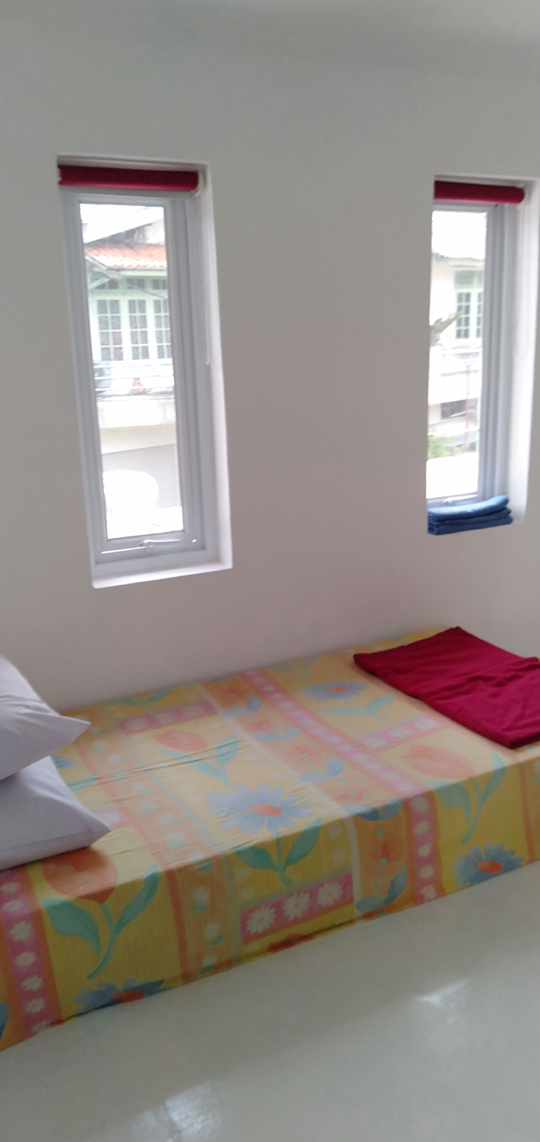 A small room with a colorful bedspread, two windows with red blinds, and a white pillow on the bed.