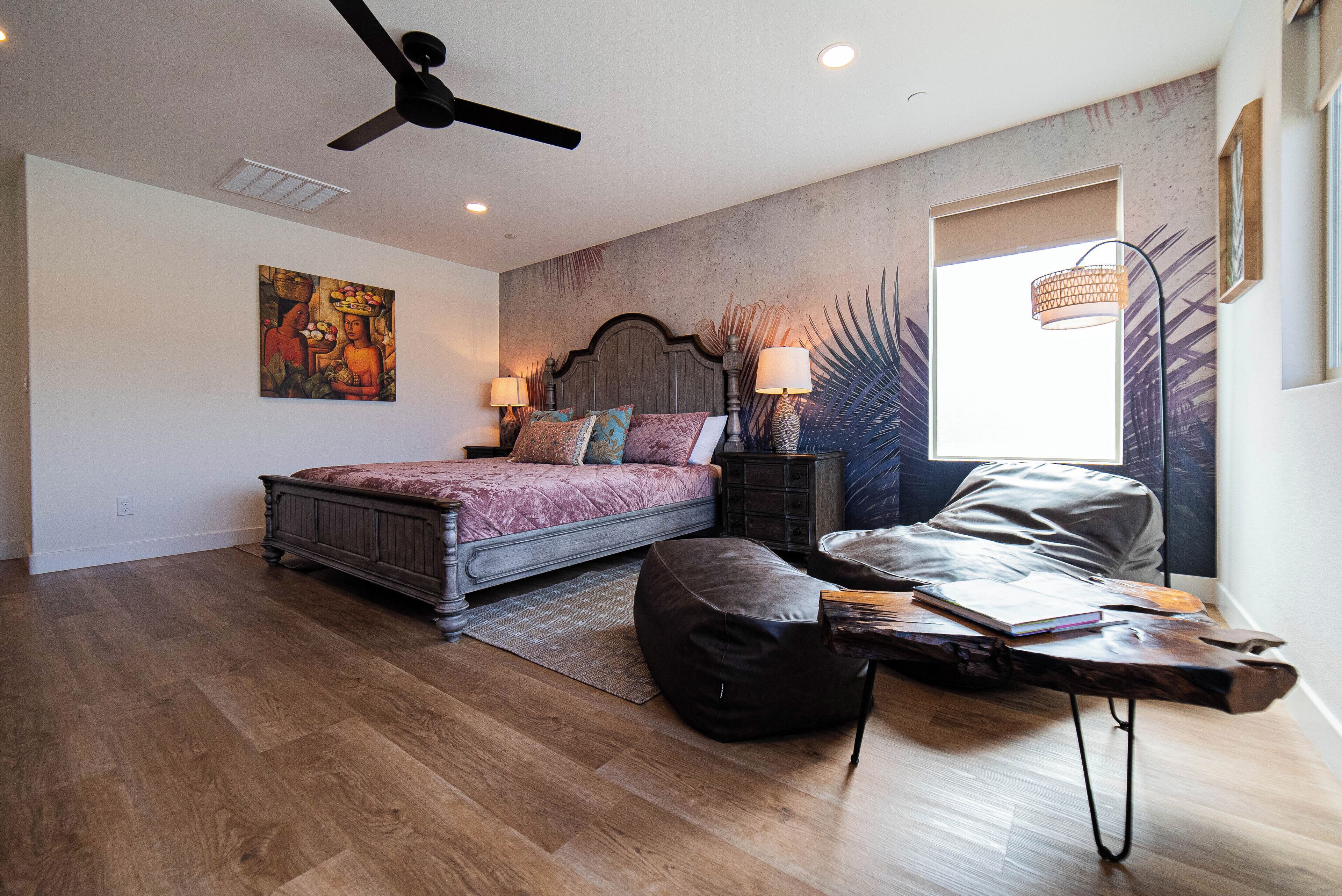 A bedroom with a bed, a desk, a chair, a ceiling fan, and a window with a plant.