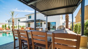 Ein überdachter Essbereich im Freien mit Holzkästen und Grill, mit Blick auf einen Swimmingpool und eine Wohngegend.