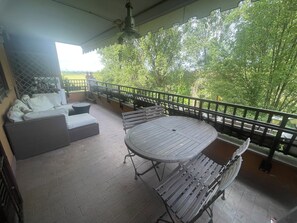 Ein überdachter Außenbereich mit Tisch und Stühlen, einer Couch und einem Geländer mit Blick auf eine grüne Landschaft.