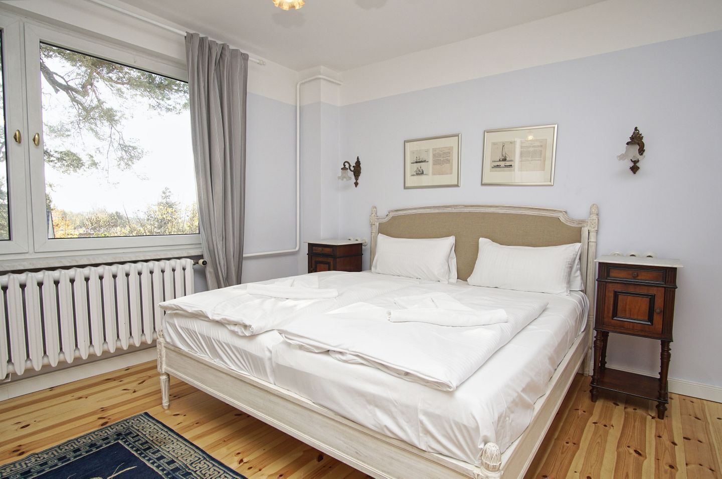 Ein ordentlich bezogenes Bett in einem Zimmer mit Heizkörper, einem fenster mit Vorhängen und zwei gerahmten Bildern an der Wand.